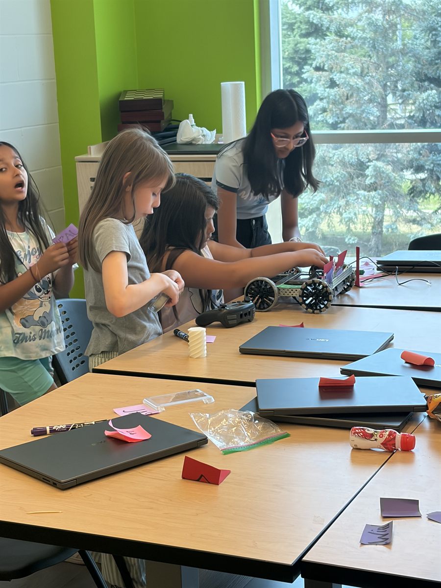 Two children building a wheeled robot with a Code Sensei looking on, laptops open on the table.