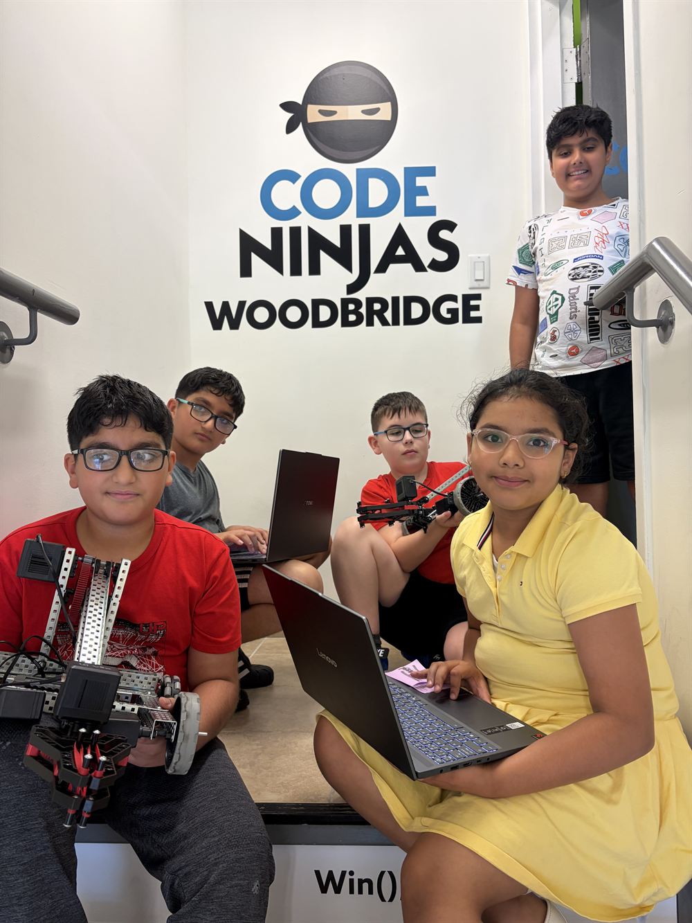 Five ninjas with their robots and laptops in front of the Code Ninjas Woodbridge wall sign.