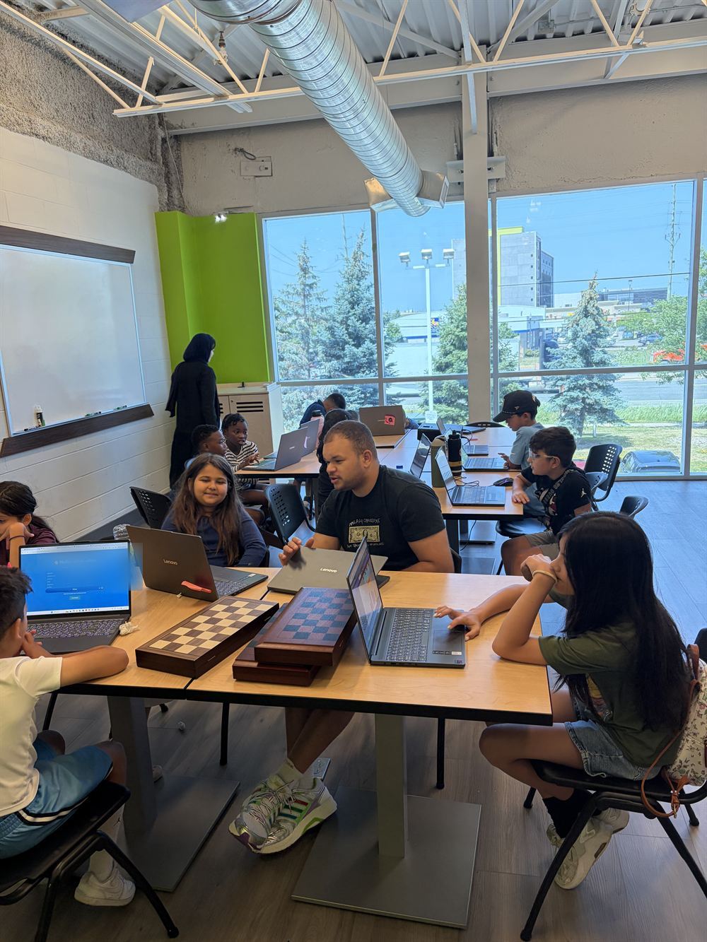 The main room at Code Ninjas Woodbridge: long tables, laptops and chess boards, a Code Sensei working with a group beside floor-to-ceiling windows.